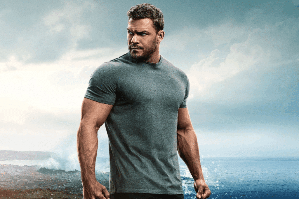 Protagonista da série "Reacher", usando camisa cinza, em pé e olhando para o lado. Céu cinza acima dele 