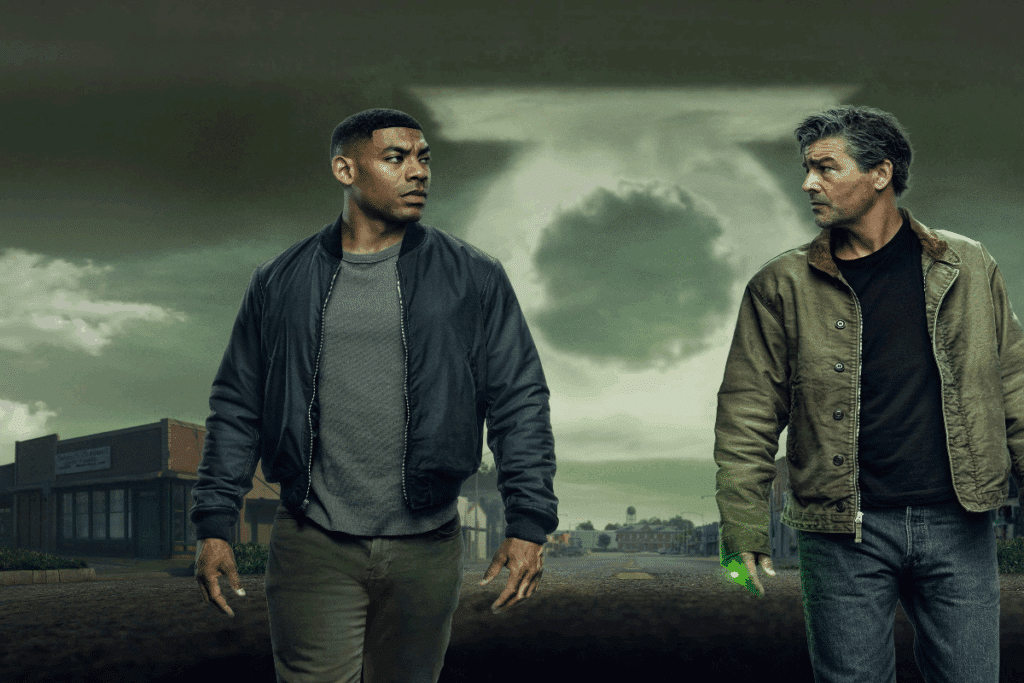 Dois homens um ao lado do outro, um deles tem o cabelo curto, e está usando camiseta e casaco cinza e calça jeans, e o outro tem o cabelo curto grisalho e está usando camiseta preta e casaco verde, e anel com luz verde. Com fumaça formando o anel do lanterna verde ao fundo e rua principal de uma cidade