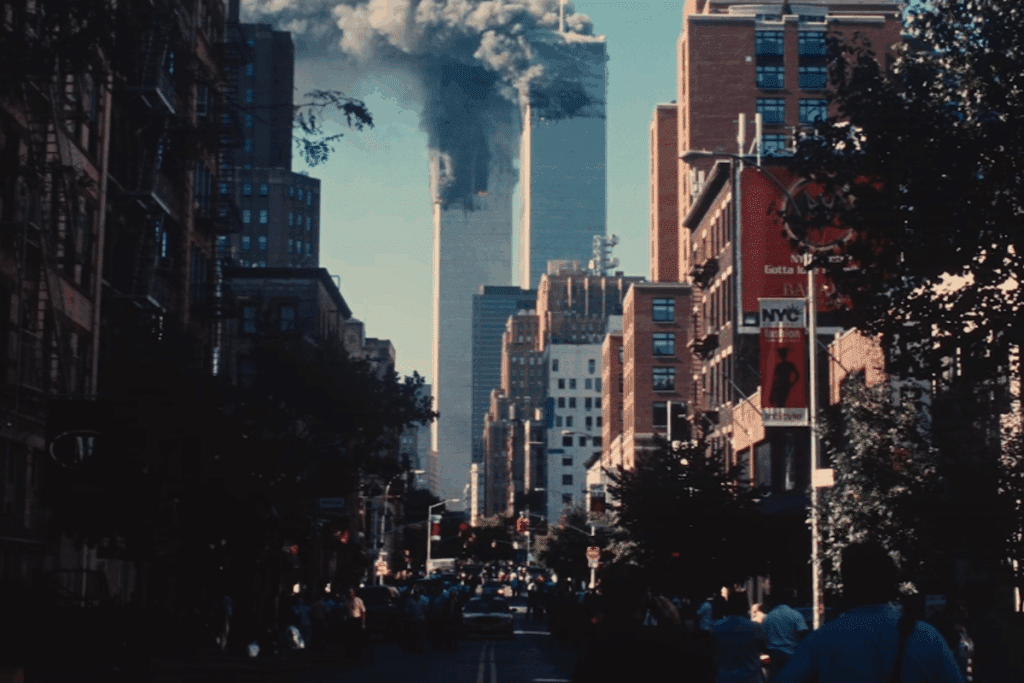 Uma rua movimentada de cidade grande com visão ao fundo das Torres Gêmeas atingidas e soltando fumaça escura no céu.