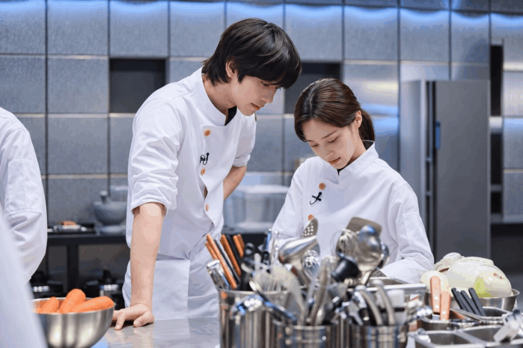 Cena de "The Final Table". Ahn Hyo-seop e Hong Hwa-yeon, com dólmãs brancos de chef, estão numa cozinha profissional de inox. Eles olham com atenção para a bancada, ao lado de recipientes cheios de utensílios, cenouras e cebolas.