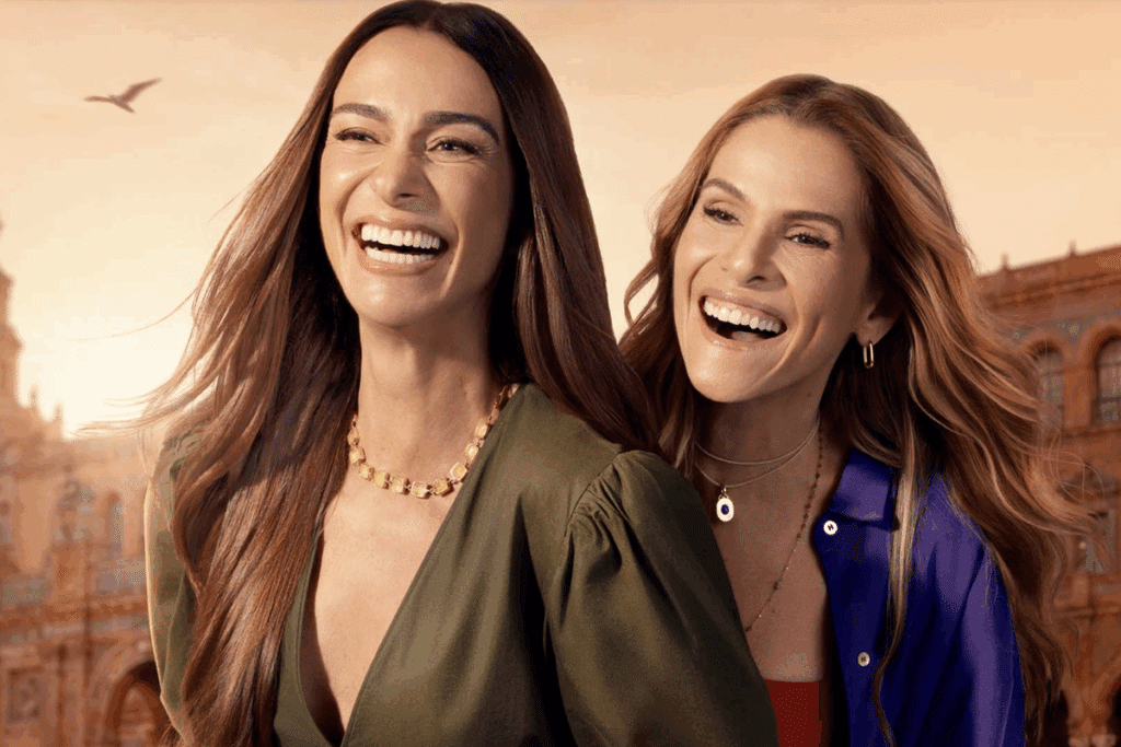 Duas mulheres sorrindo abraçadas ao ar livre, uma de vestido verde-oliva com as mãos no guidão de uma patinete e a outra de camisa azul apoiada em seu ombro, tendo ao fundo um edifício histórico sob céu alaranjado.