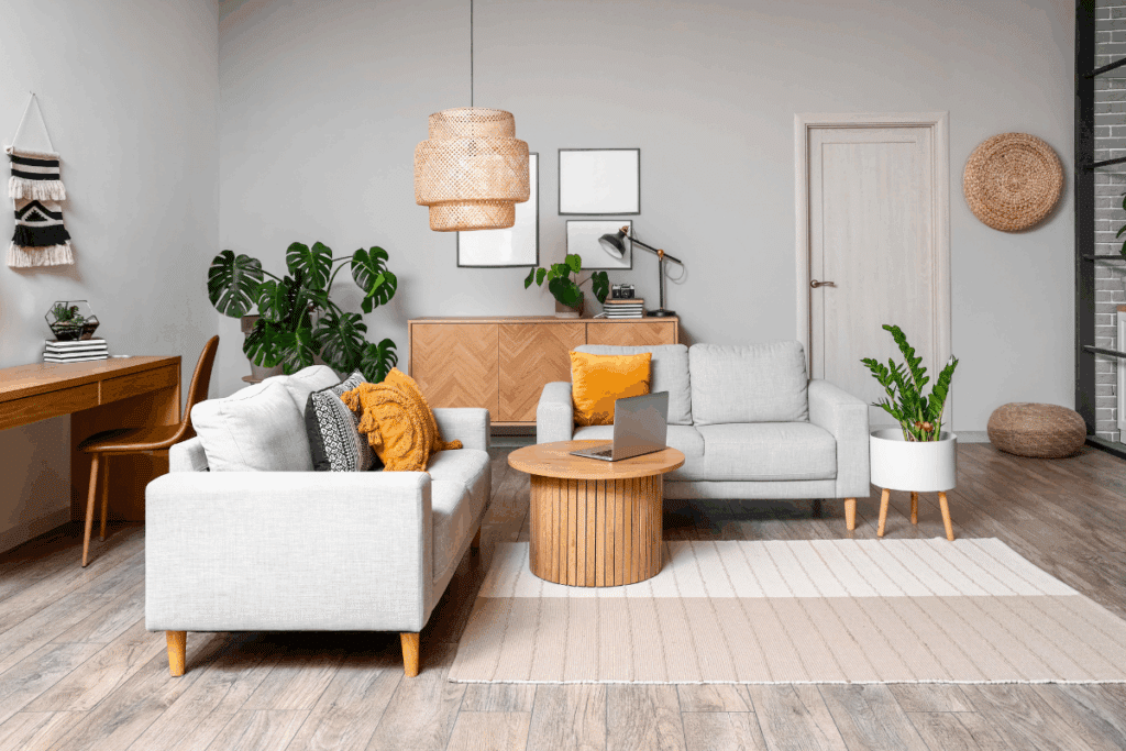 Sala de estar com dois sofás cinza-claro, almofadas brancas, pretas e amarelas e mesa de centro redonda de madeira com um notebook aberto. Ao fundo, há um aparador de madeira clara, plantas verdes, quadros minimalistas e uma luminária pendente de fibras naturais. À esquerda, uma escrivaninha de madeira com cadeira marrom. O ambiente tem paredes brancas, piso amadeirado e tapete bege.