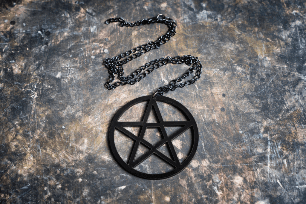 Fotografia de um colar com pingente de pentagrama preto, uma estrela de cinco pontas com uma ponta voltada para cima dentro de um círculo, preso a uma corrente escura. O colar está sobre uma superfície envelhecida com manchas em tons de cinza, marrom e dourado.